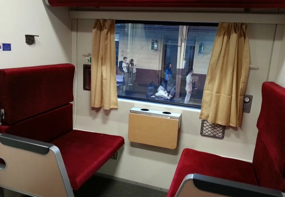 The interior of a train cabin in Thailand featuring red upholstered seats, a small wooden fold-down table, and a window view of the platform.