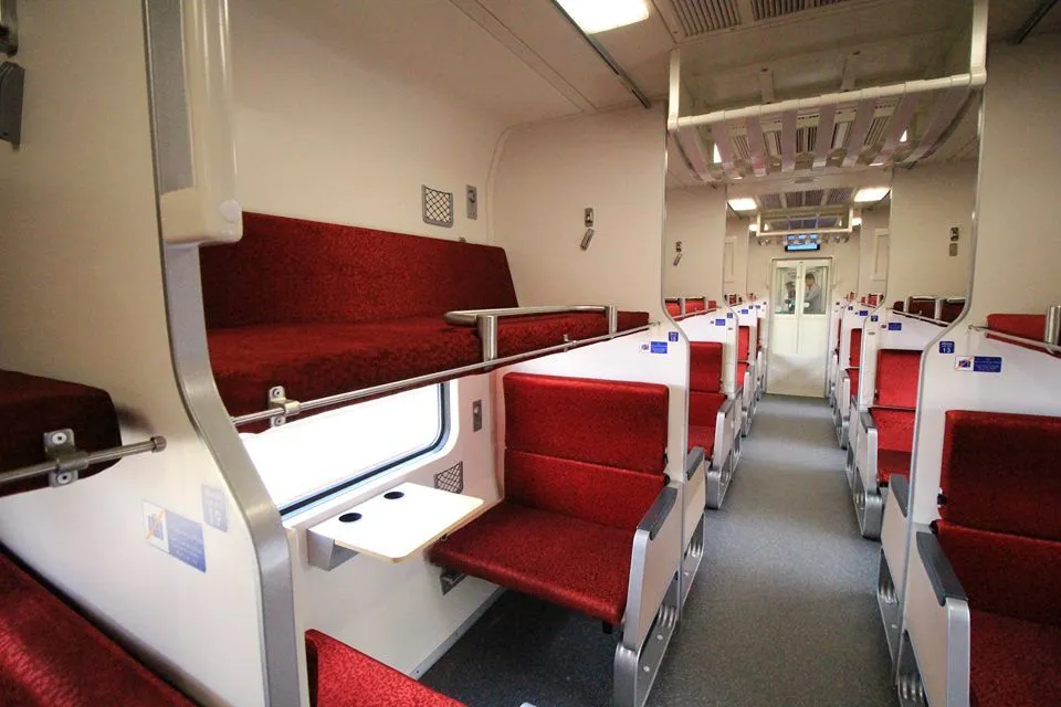 The interior of a train cabin in Thailand featuring red upholstered seats, a small wooden fold-down table, and a window view of the platform.