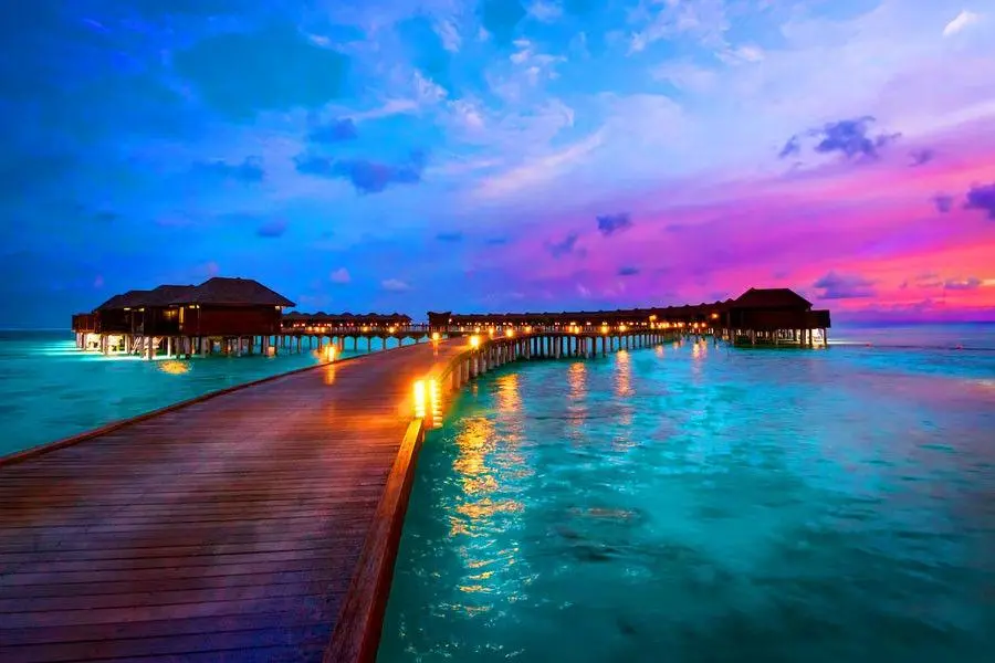 A wooden walkway leads to overwater bungalows in the Maldives under a vibrant sunset sky, with lights reflecting on the turquoise water.