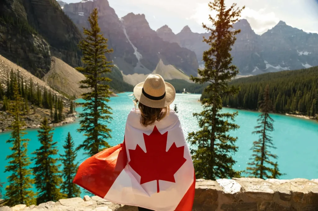 A woman wearing a sun hat and draped in a Canadian flag looks out at the vibrant turquoise waters of Moraine Lake surrounded by mountains.