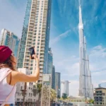 A woman wearing a red headscarf uses her smartphone to take a photo of the iconic Burj Khalifa skyscraper in Dubai on a bright, sunny day.