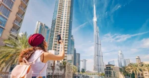 A woman wearing a red headscarf uses her smartphone to take a photo of the iconic Burj Khalifa skyscraper in Dubai on a bright, sunny day.
