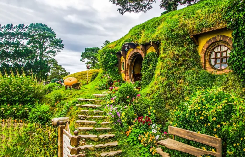 A cozy, grass-covered Hobbit hole with a round wooden door and window, nestled into a green hillside with a stone path and vibrant flowers.