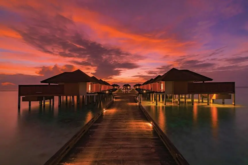 A wooden walkway leads to overwater bungalows in the Maldives under a vibrant sunset sky, with lights reflecting on the turquoise water.