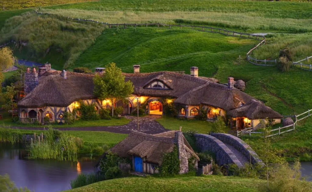 The charming Hobbiton village in New Zealand features cozy thatched-roof houses and a stone bridge over a pond, illuminated by warm lights at dusk.