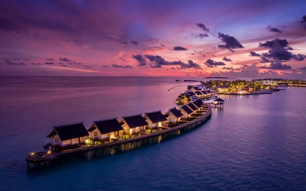 A wooden walkway leads to overwater bungalows in the Maldives under a vibrant sunset sky, with lights reflecting on the turquoise water.