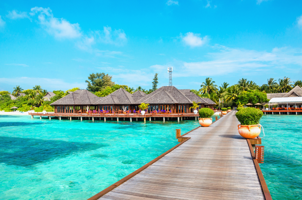 A wooden walkway leads to a restaurant built over turquoise water at a tropical island resort, surrounded by palm trees and a blue sky.