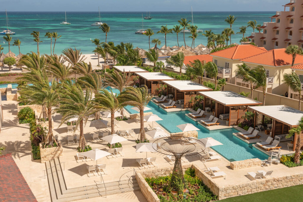 Aerial view of a luxury tropical resort pool with palm trees, sun loungers, cabanas, and the ocean with sailboats in the background