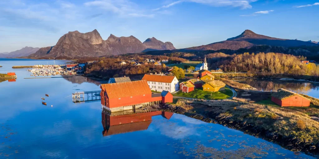 An aerial view of a bright red traditional Norwegian rorbu cabin sitting directly on the water's edge, reflecting in a serene fjord with dramatic mountain peaks under a clear blue sky.