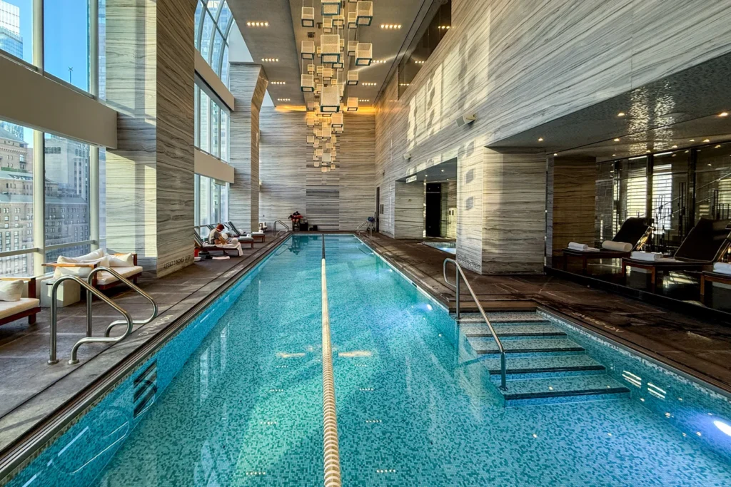 An indoor lap pool with blue mosaic tiles, large windows, lounge chairs, and modern hanging light fixtures in a luxury hotel