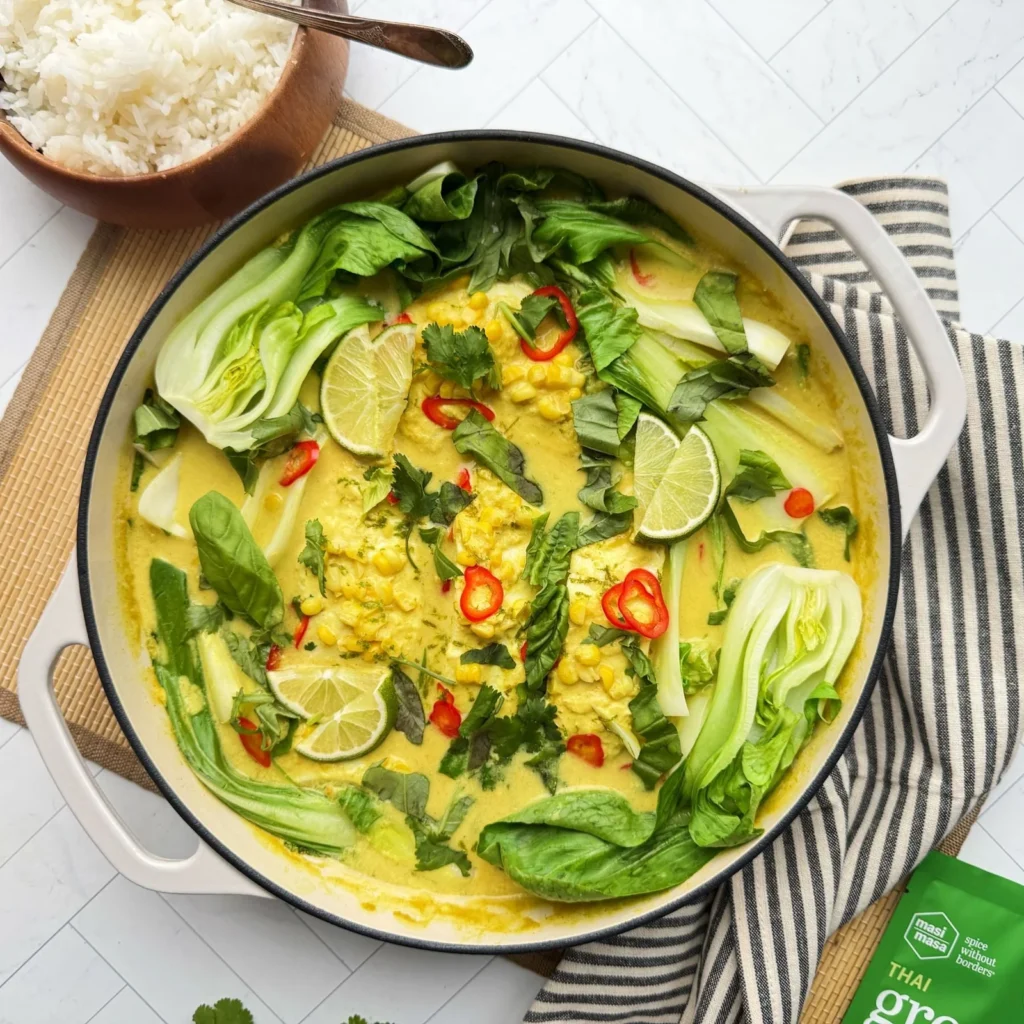 A skillet of creamy Thai green curry with cod, corn, bok choy, and lime slices, served with a bowl of rice.