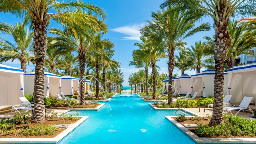 Long swimming pool at Grand Hyatt Baha Mar surrounded by palm trees, water fountain arcs, and private luxury cabanas under a bright blue sky.
