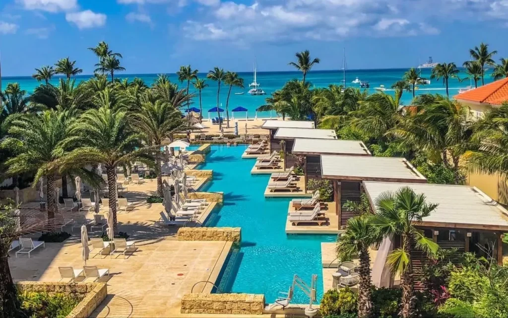 Aerial view of a luxury resort swimming pool surrounded by palm trees and cabanas, leading out to a tropical white sand beach and turquoise sea.