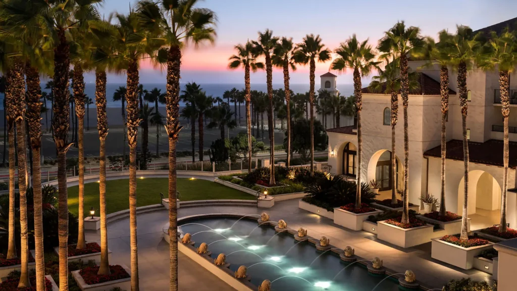 Evening view of Hyatt Regency Huntington Beach Resort featuring an illuminated courtyard fountain, tall palm trees, and ocean views.