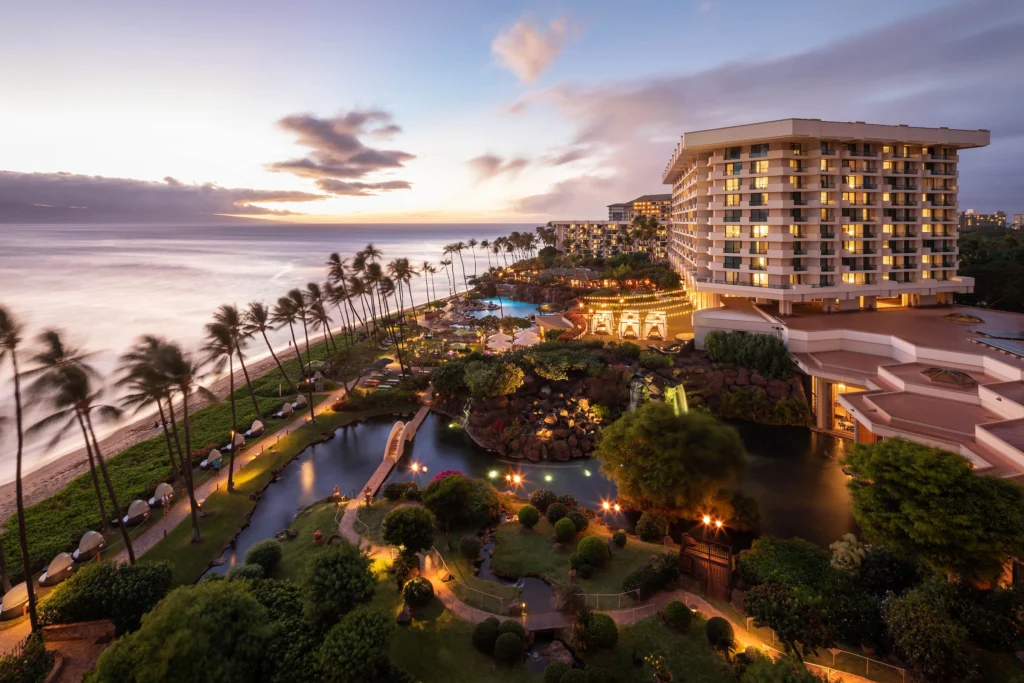 Elevated twilight view of a grand beachfront resort hotel with glowing lights, manicured gardens, palm trees, and a pathway running alongside the ocean at sunset.