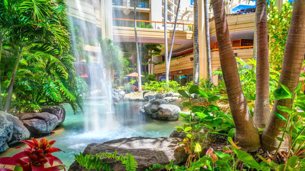 Open-air atrium at Hyatt Regency Waikiki Beach Resort featuring a cascading waterfall, lush greenery, and surrounding boutique shops.