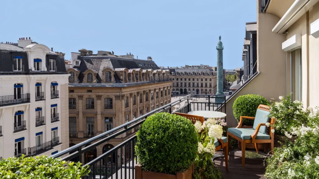 A hotel balcony decorated with potted plants and seating, offering a view of classic Parisian architecture and a tall column in the square