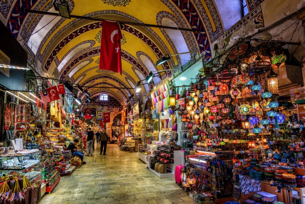 Inside the bustling Grand Bazaar in Istanbul, featuring vaulted ceilings with Turkish flags and shops selling colorful mosaic lamps and souvenirs