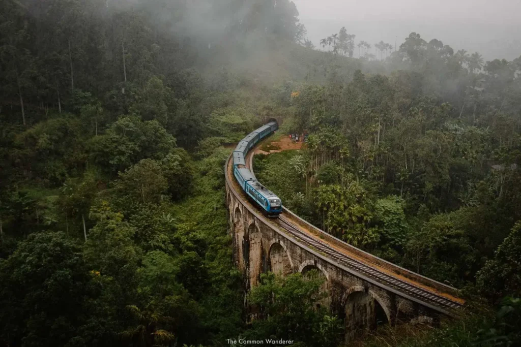 A blue train crossing the iconic Nine Arch Bridge surrounded by lush green misty forests and hills in Sri Lanka.