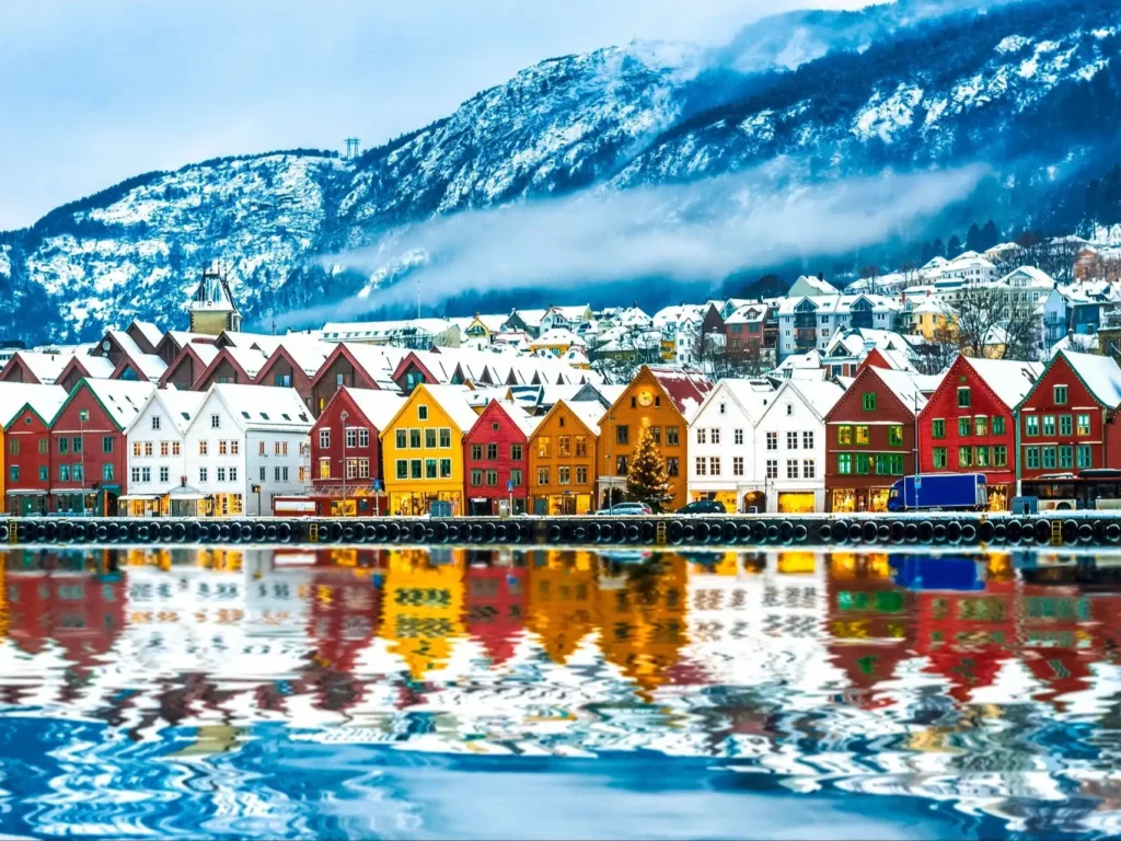 A scenic winter view of traditional colorful wooden buildings along the waterfront in Bergen, Norway, with snow-covered rooftops and mountains in the background reflecting brightly in the calm water.