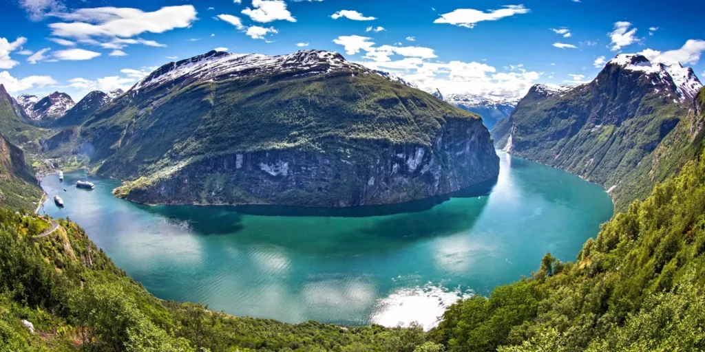 A wide panoramic landscape shot of the deep turquoise Geirangerfjord curving between dramatic steep cliffs and snow-capped mountains under a bright blue sky.