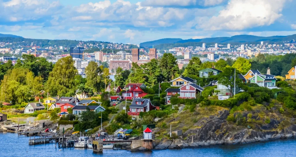 A picturesque view of colorful wooden houses nestled on a lush, rocky island shoreline with a small dock, overlooking the waters and city skyline of Oslo in the background.