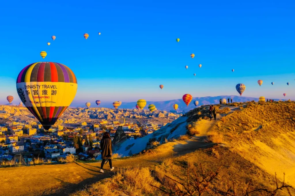 A traveler overlooking a landscape filled with colorful hot air balloons rising above the town and valleys of Cappadocia in Turkey.