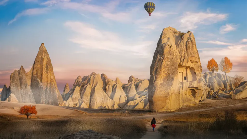 A lone traveler walking past unique rock formations and carved fairy chimneys in Cappadocia under a sky with a hot air balloon