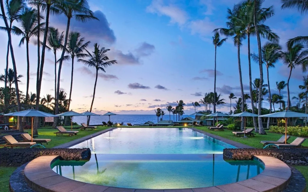 Evening view of the infinity pool at Hana-Maui Resort surrounded by palm trees, lounge chairs, and umbrellas overlooking the ocean at sunset