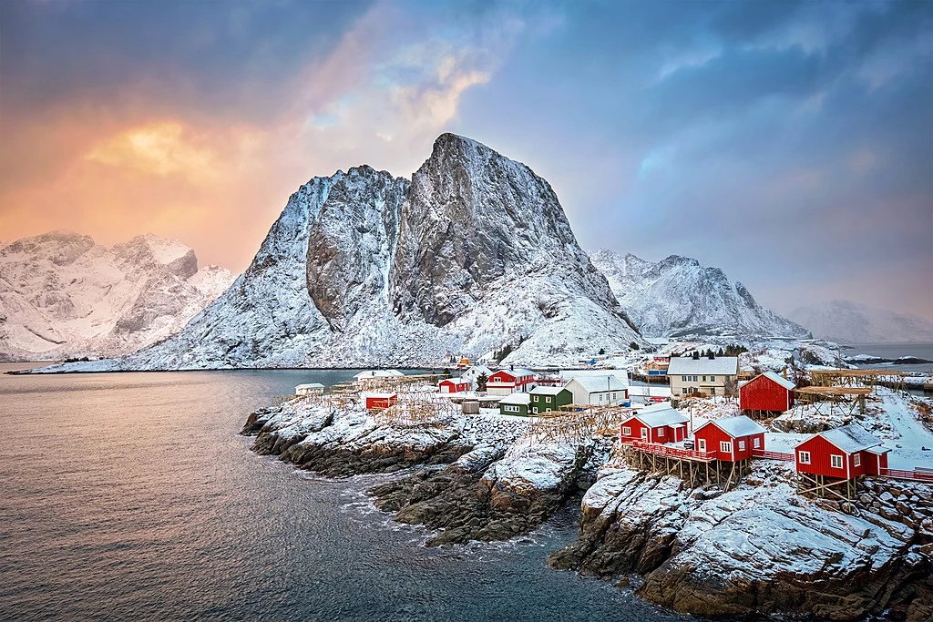 A dramatic winter landscape featuring iconic red rorbu cabins built on snow-covered rocky shores, set against a majestic snow-capped mountain peak under a glowing sunset sky in Lofoten, Norway.