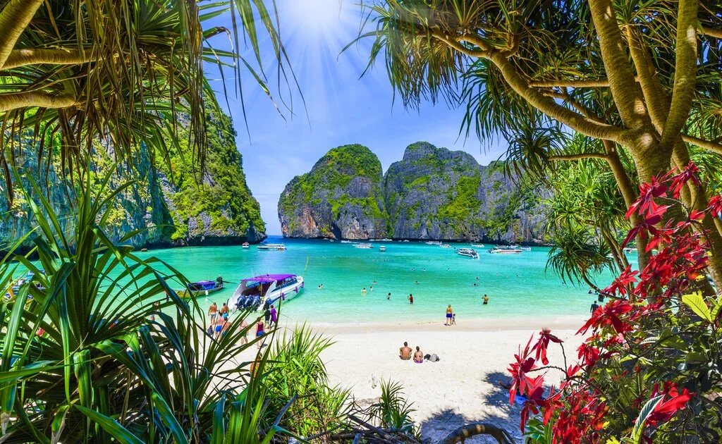 A picturesque tropical beach framed by lush green trees and red flowers, overlooking a turquoise bay with boats and limestone cliffs.