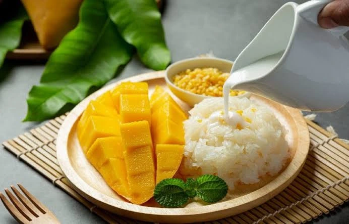 Sweet sticky rice served with fresh yellow mango slices on a wooden plate, with coconut milk being poured from a pitcher.