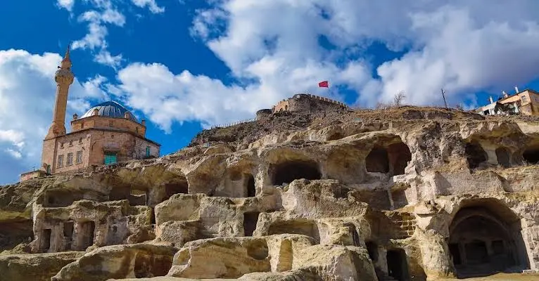 Carved ancient rock caves and dwellings in Cappadocia featuring a historic stone mosque with a minaret against a cloudy blue sky