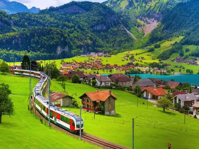 A red and white scenic train winding along tracks through a lush green valley, passing by a traditional Swiss village with red-roofed houses against a backdrop of towering mountains.