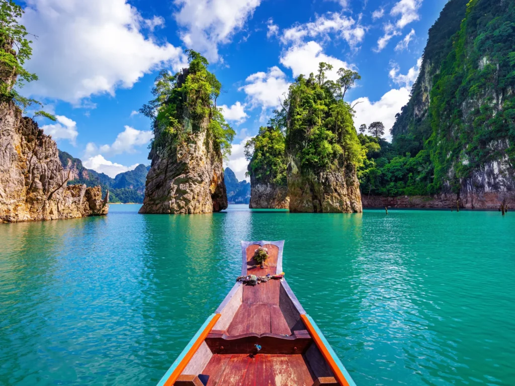 View from the front of a traditional wooden long-tail boat sailing through turquoise waters surrounded by towering limestone karsts in Thailand