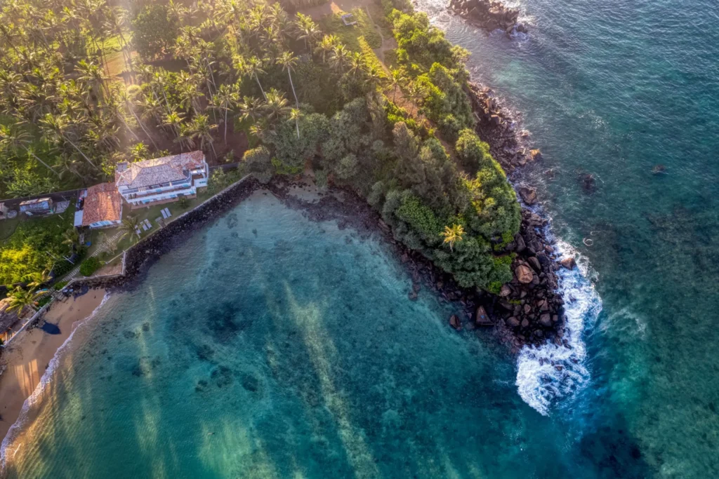 Aerial view of a scenic tropical coastline featuring a sandy beach, clear blue water, rocky point, and palm trees in Sri Lanka.