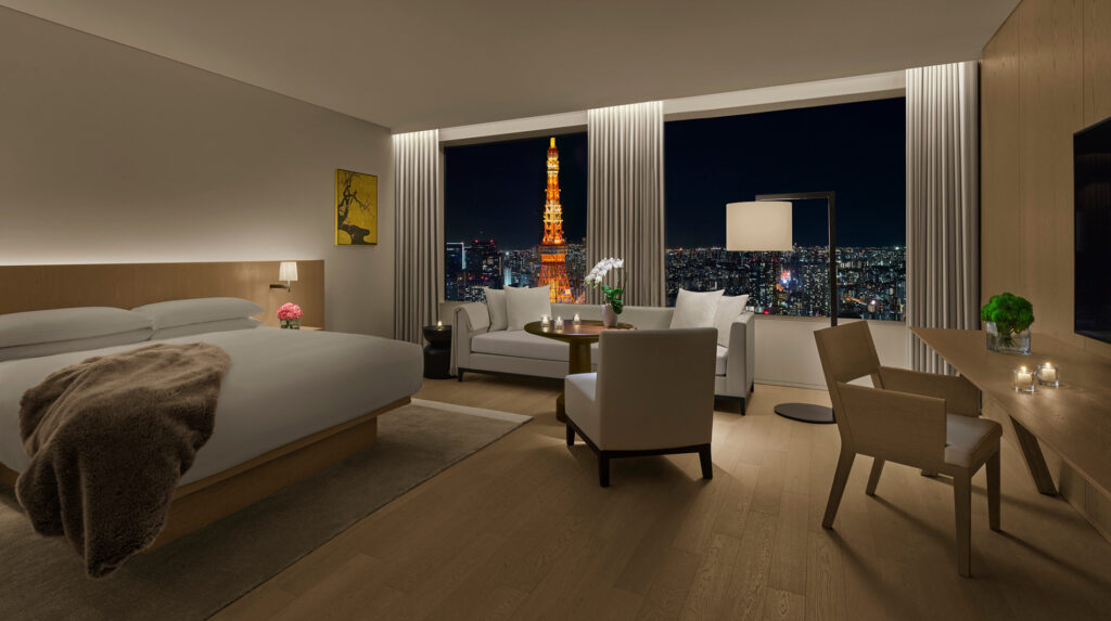 Luxury hotel suite bedroom featuring a king bed, sitting area, desk, and large windows with a night view of the illuminated Tokyo Tower