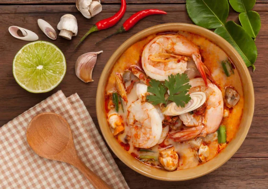 A bowl of spicy Thai Tom Yum Goong soup with shrimp, garnished with herbs, and surrounded by ingredients like lime, chili peppers, and lemongrass on a wooden table.