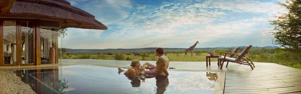 A couple relaxing and drinking wine in an infinity pool on a lodge deck while viewing wildlife on the African savanna.