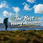 Two hikers with backpacks and trekking poles walking along a grassy ridge overlooking a lake and distant mountain ranges