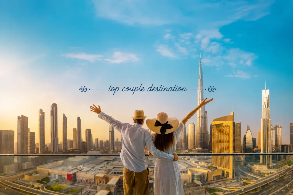 A couple standing with arms raised on a balcony overlooking the modern Dubai city skyline and Burj Khalifa.