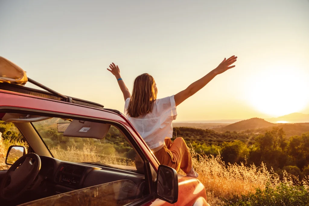 A woman sitting on the hood of a red car with her arms raised wide, enjoying the sunset view over a landscape