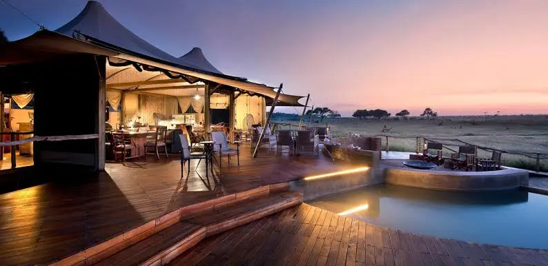 A luxury safari camp deck with a swimming pool and open tent lounge overlooking the African savanna at sunset.