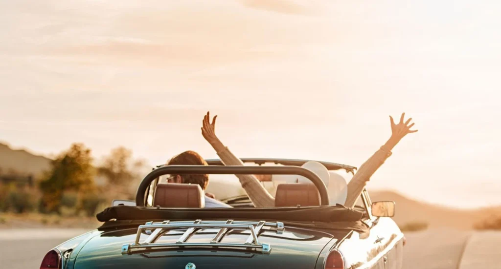 A couple with raised arms driving a convertible car on an open road during sunset